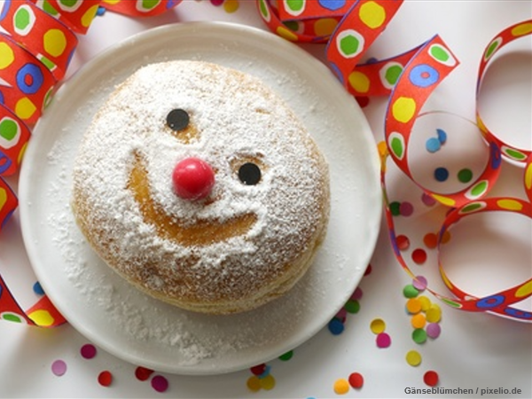 Ein süßes Gebäck (Berliner) verziert mit einem lächelndem Gesicht. Im Hintergrund Luftschlange und Konfettin